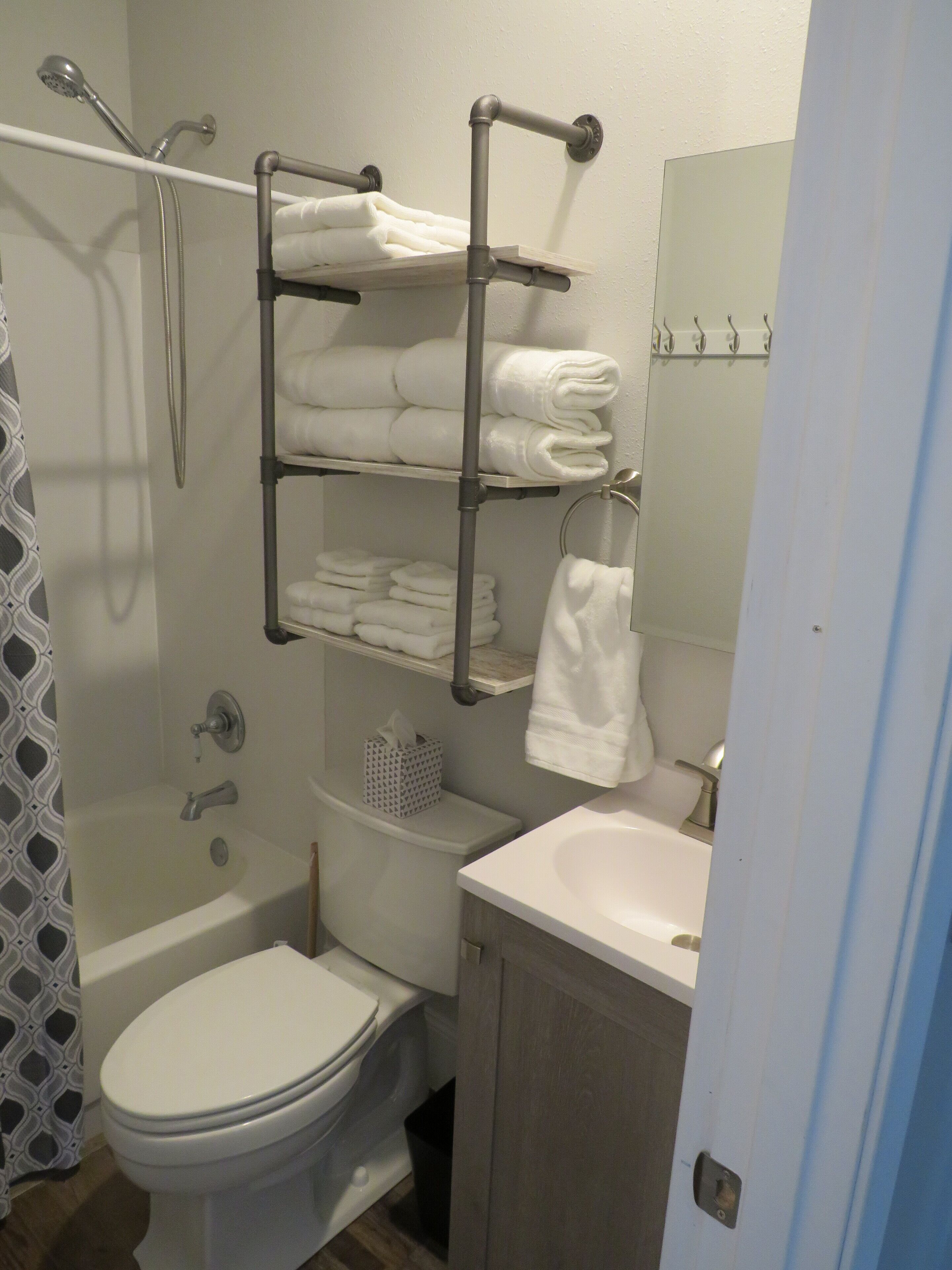 A bathroom with a toilet, sink, and shower. There are shelves with towels and a tissue box.