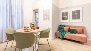 Een zithoek met een ronde tafel, groene stoelen en een bank met een groene plaid. Er hangen afbeeldingen in lijsten aan de muur en er is een raam met gordijnen.