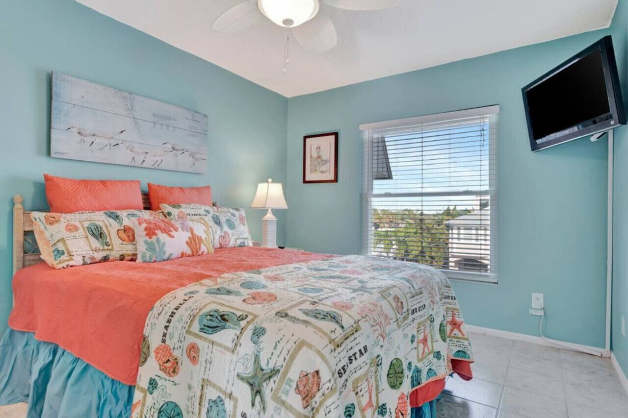 A bedroom with a bed, a ceiling fan, a TV mounted on the wall, and a window with blinds.