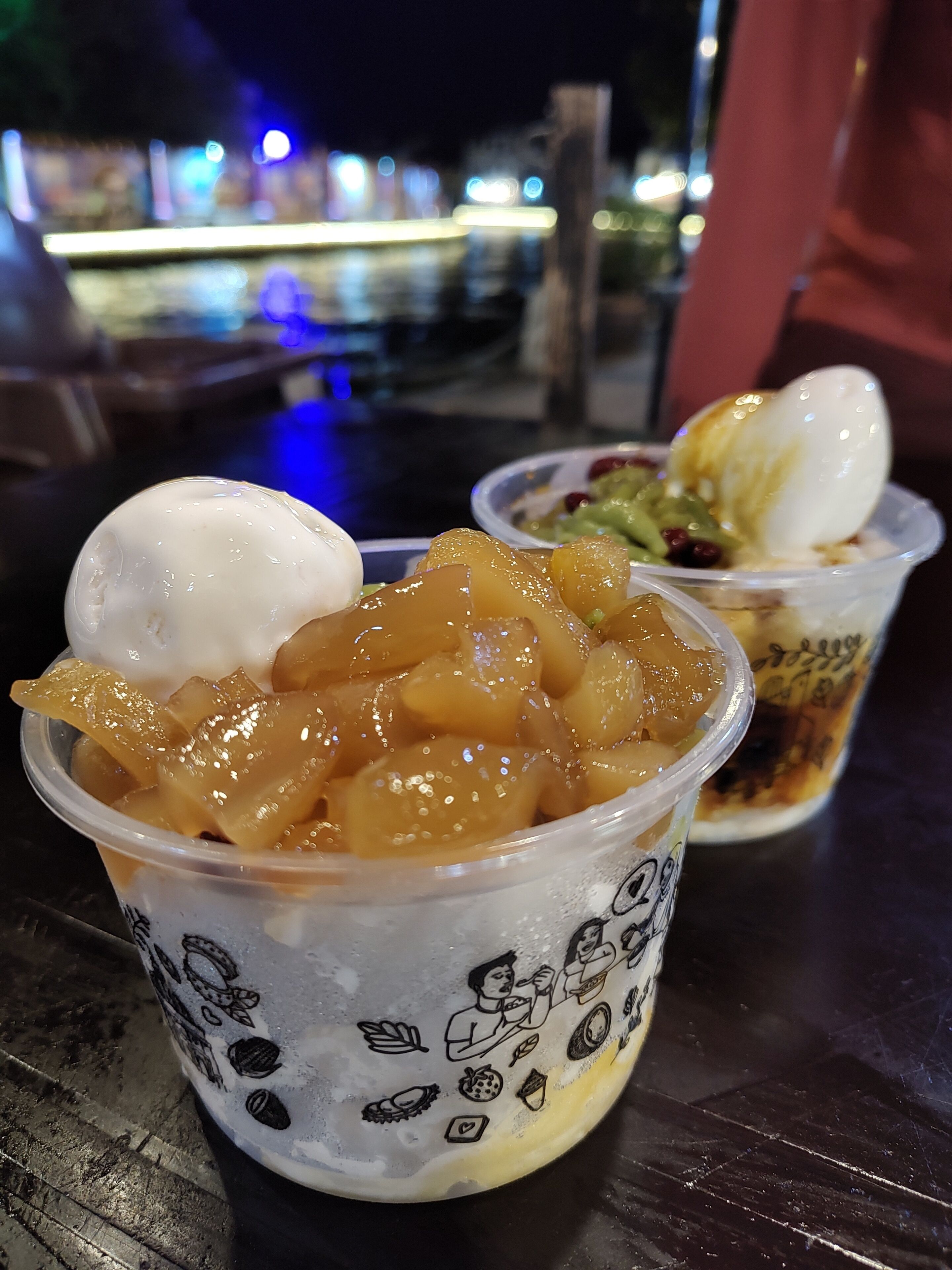 Two cups of dessert with fruit and ice cream, placed on a table with a blurred background.