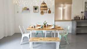 A dining area with a wooden table, white chairs, and a bench. There are string lights on the wall, a hanging lamp, and a window with blinds. - Cloth Art Homestay near Jonker&Pahlawan (Malacca City)
