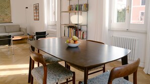 Un coin repas avec une table en bois, quatre chaises, un saladier de fruits, une bibliothèque et un radiateur.