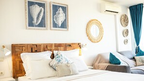 A bedroom with a wooden headboard, white bedding, and decorative wall art including a mirror and framed shells. - Cosy studio, beach access (Le Gosier)