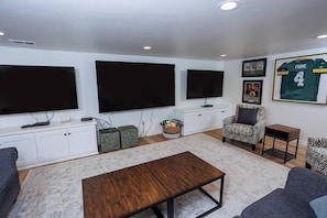 A living room with a sofa, a coffee table, a patterned armchair, a small side table, and a TV wall-mounted above a white cabinet.