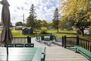 A deck with outdoor seating, a fire pit, and a view of a park with trees and a building in the background. - Cheese Head Hideaway - Walk to Lambeau Field (Green Bay)