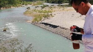 Un hombre de pie junto a un río, con montañas al fondo.