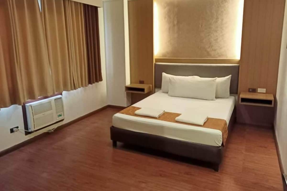 A hotel room with a large bed, a headboard with integrated lighting, a bedside table, and a wall-mounted air conditioning unit.