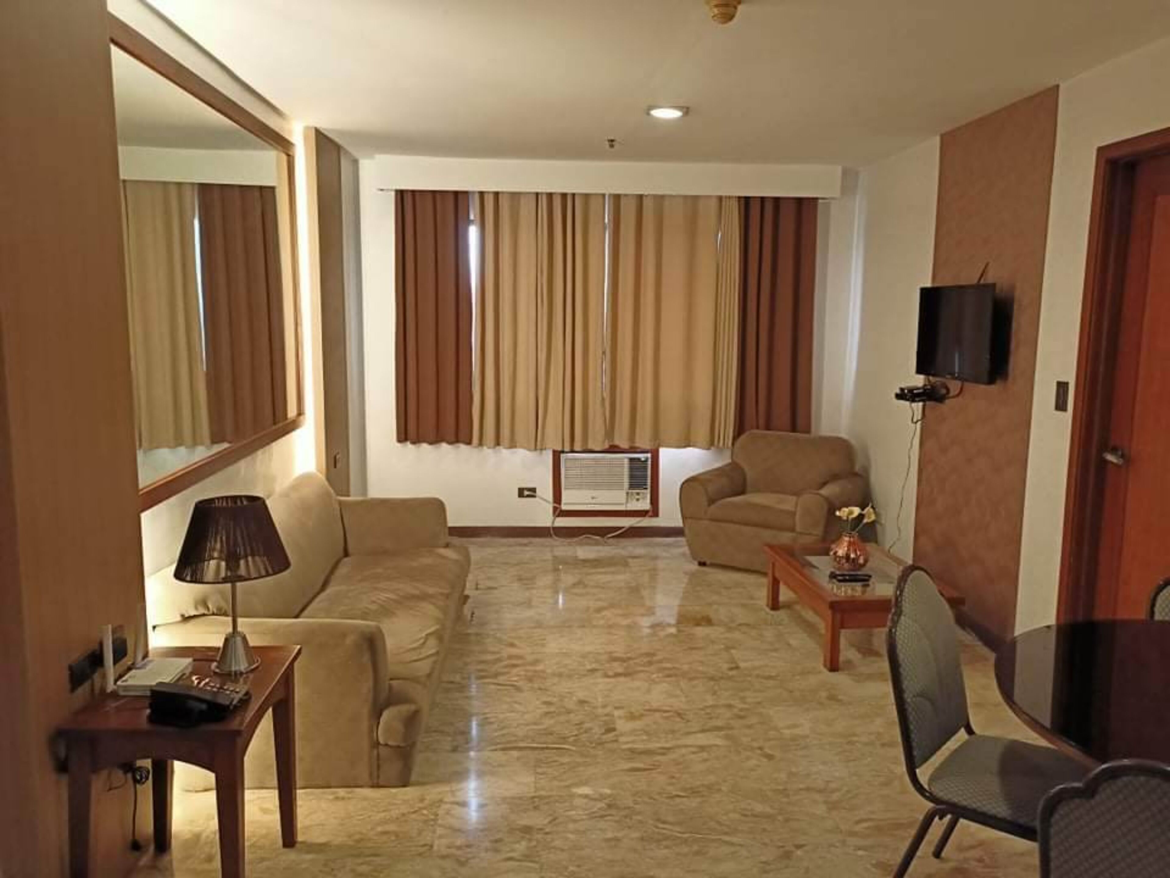 A living room with a sofa, armchair, and a small table with a lamp. There is a television mounted on the wall and a window with curtains.