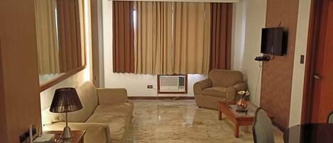 A living room with a sofa, armchair, and a small table with a lamp. There is a television mounted on the wall and a window with curtains.