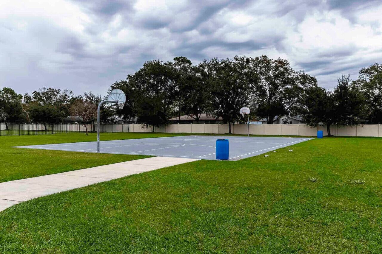 An outdoor basketball court with a fence, trees, and a cloudy sky.