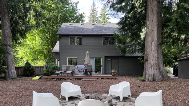 Ein Garten mit einer Steinterrasse, vier weißen Stühlen, einem überdachten Grill und einem grauen Haus mit einer Veranda.