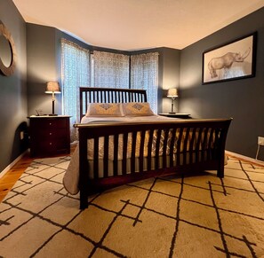 Une chambre à coucher avec un lit en bois, deux tables de chevet avec des lampes, des rideaux à motifs et un tableau encadré au mur.