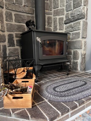 Una estufa de leña con chimenea, un caja para guardar leña y una alfombra estampada sobre un suelo de piedra.