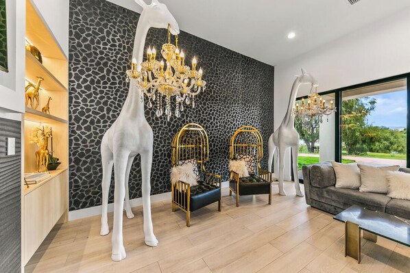 A modern living room with a giraffe sculpture, two golden chairs, a chandelier, and a sofa with a view of the outdoors.