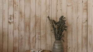 Un mur en bois rustique orné d’une ampoule suspendue, une petite étagère en bois avec des livres et un vase de fleurs séchées, et un banc avec un coussin.