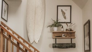 Un escalier avec des rampes en bois, une planche de surf, une étagère murale agrémentée de plantes et d’objets décoratifs, et des tableaux encadrés.