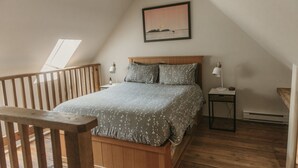 Une chambre avec un plafond en pente, un cadre de lit en bois, une couvre-lit à motifs floraux gris, deux lampes de chevet, une petite table de chevet et un tableau encadré accroché au mur.