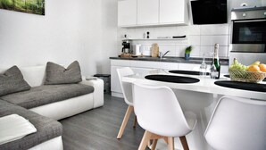 Eine moderne Küche mit weißen Schränken, ein Essbereich mit weißen Stühlen und ein graues Sofa.