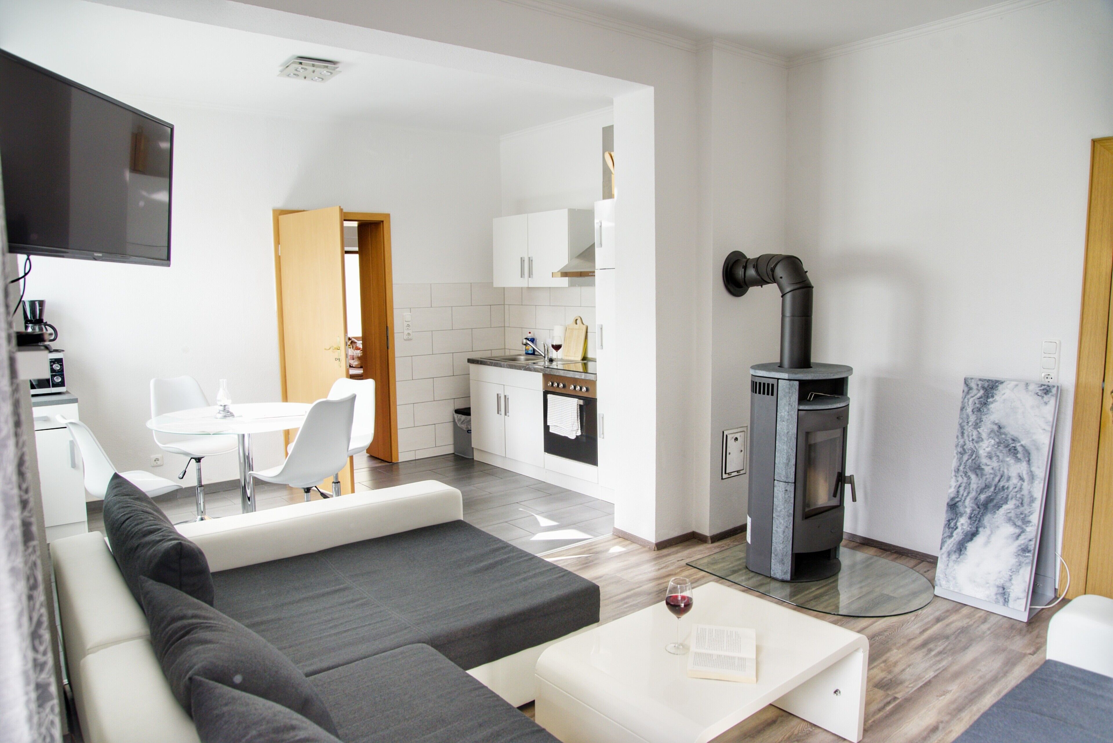 A modern living room with a sofa, a coffee table with a book and wine glass, a TV, and a kitchen with a stove and cabinets.