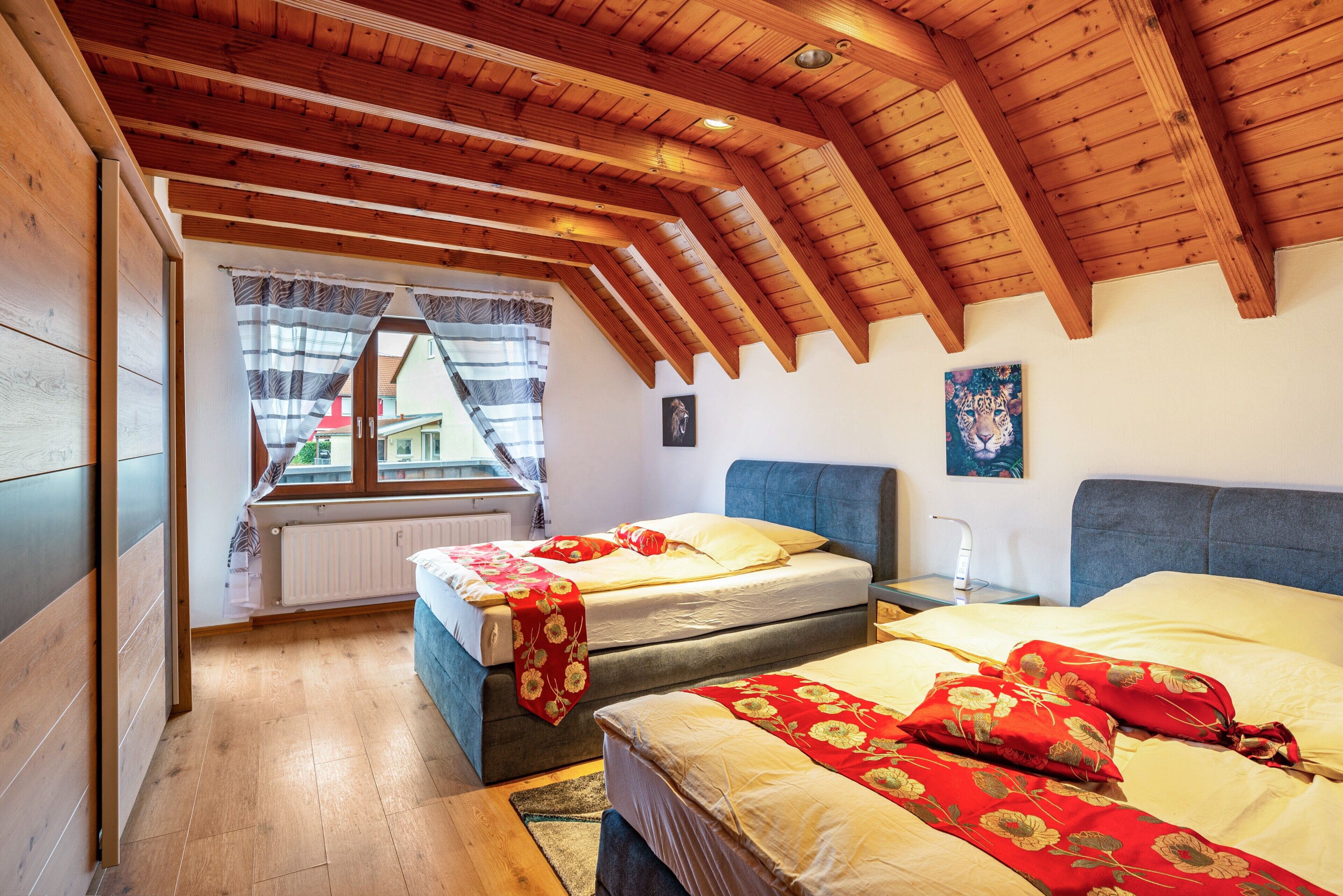 A bedroom with two beds, wooden ceiling beams, a window with curtains, and a radiator.