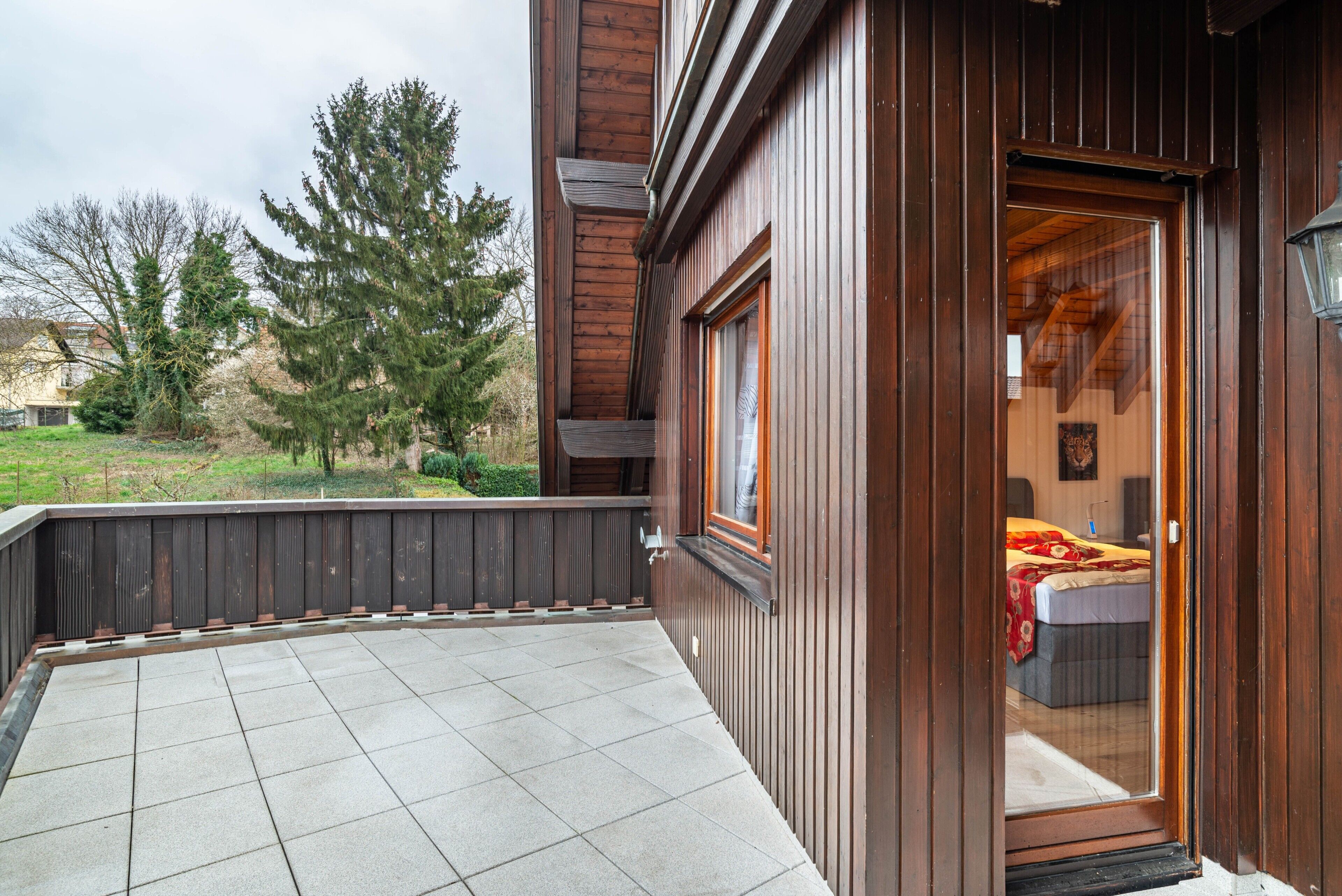 Ein Holzbalkonbau mit einem Balkon, ein durch eine Glastür sichtbares Schlafzimmer und Blick auf Bäume sowie eine Rasenfläche.