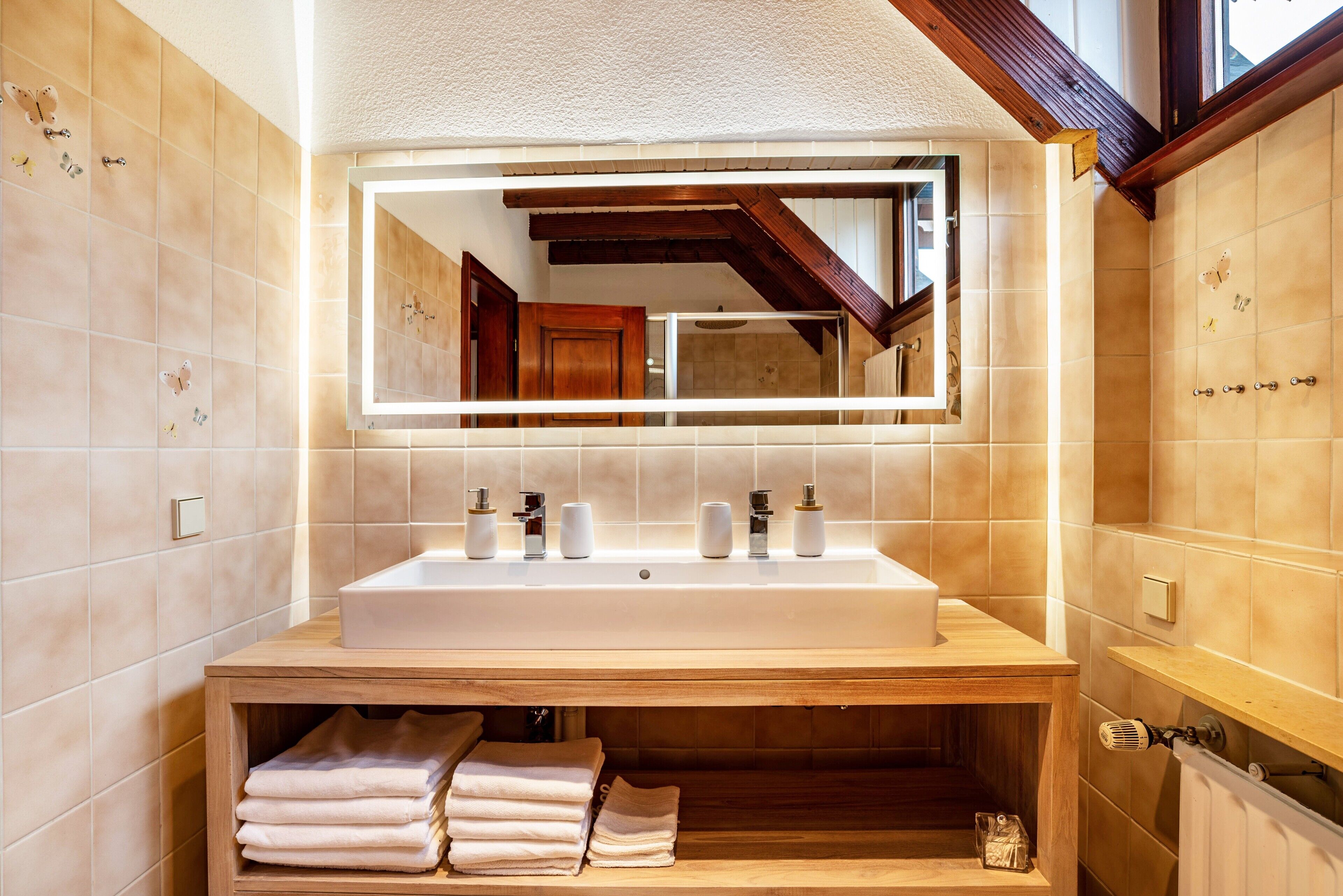 Un bagno con uno specchio grande, un lavabo con due rubinetti e un mobile contenitore in legno con asciugamani.