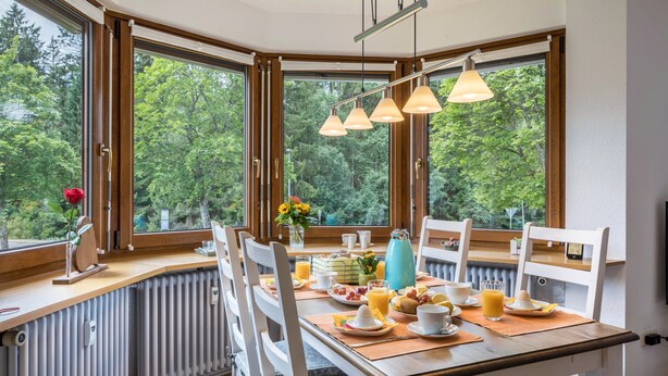 Une table de petit-déjeuner placée devant de grandes fenêtres donnant sur les arbres, avec un radiateur et une chaise blanche.