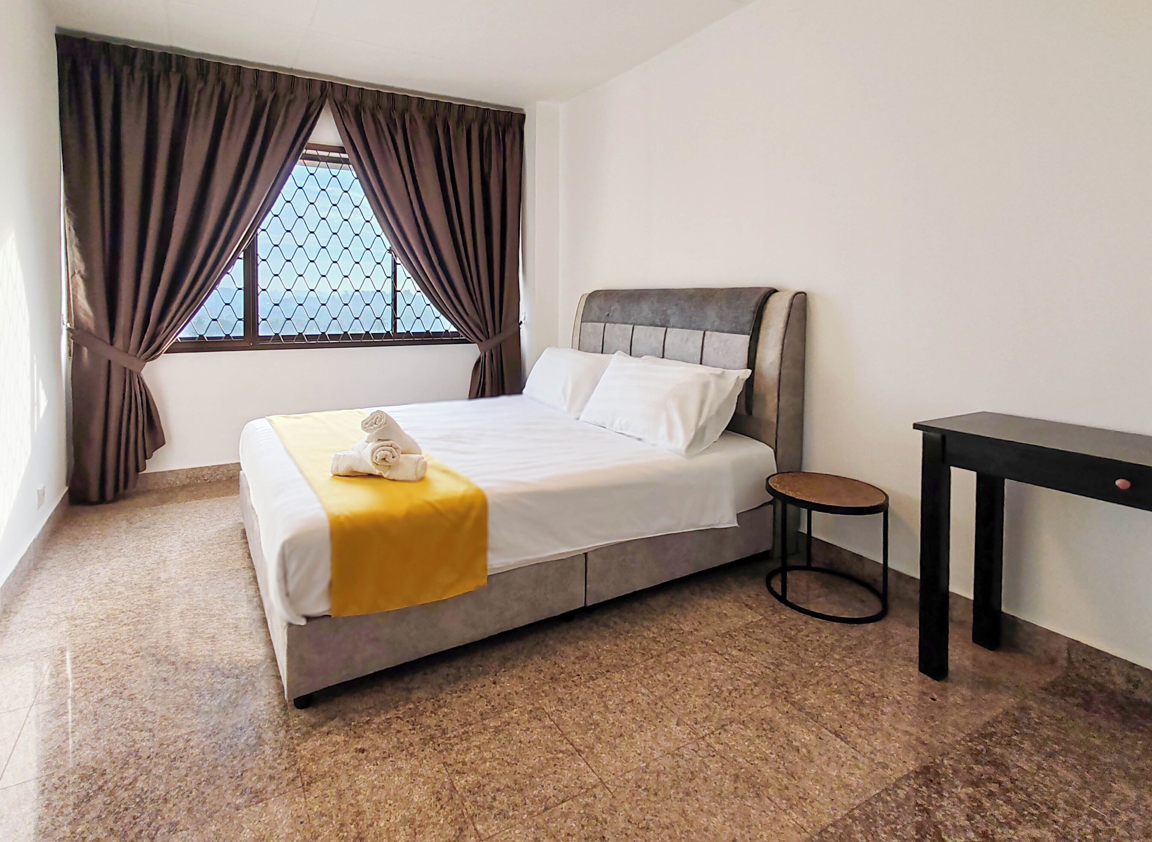 Una cama bien hecha con una colcha amarilla y una toalla enrollada, una pequeña mesa redonda a su lado y una ventana con cortinas.