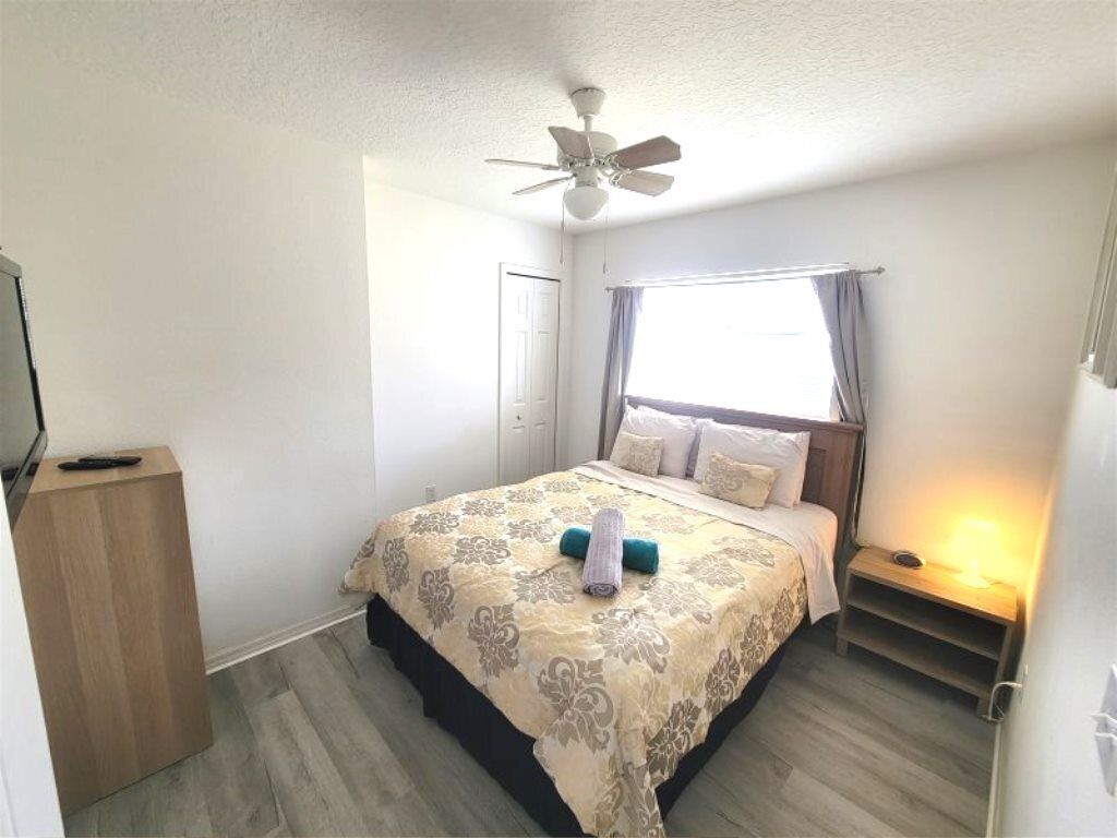 A bedroom with a bed, a ceiling fan, a bedside table with a lamp, and a window with curtains.