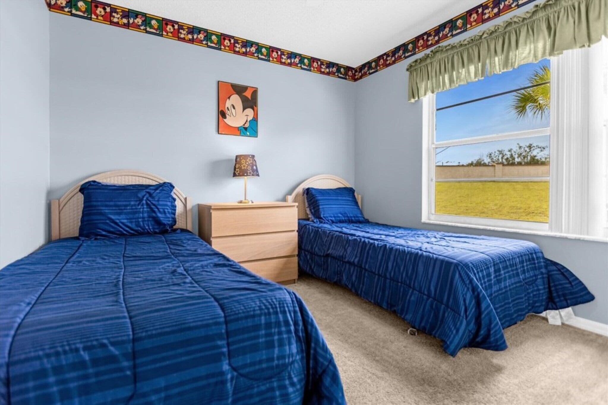 A bedroom with two beds, a dresser, a lamp, and a window with a view of a grassy field.