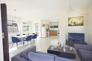 Un salon moderne avec un canapé, une table basse et un téléviseur. On y trouve également un coin repas avec des chaises et une table. La cuisine est visible au fond.