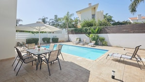 Un área de piscina con sillones de descanso, una mesa con sillas y una sombrilla blanca.