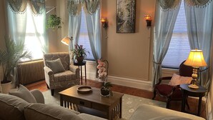 Una sala de estar con un sofá, un sillón, una mesa de centro y dos lámparas. Hay cortinas, un cuadro y vistas al exterior.