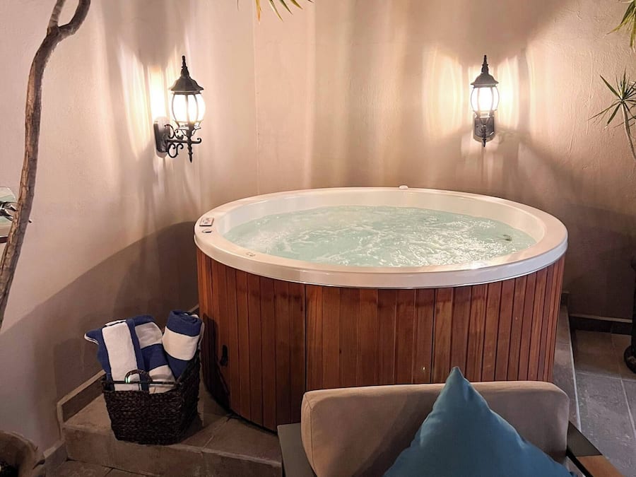 Ein Whirlpool mit Holzverkleidung, zwei Wandleuchten, ein Sessel mit Kissen und ein Korb mit Handtüchern.