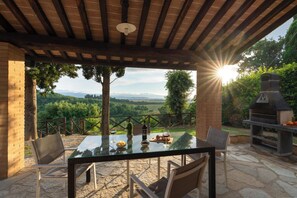 An outdoor patio with a glass table, chairs, a brick oven, and a scenic view.