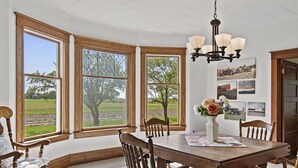 A dining area with a wooden table, chairs, and a chandelier. Large windows with wooden frames offer views of a green landscape.