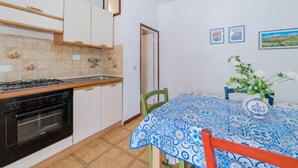 A kitchen with a stove, oven, and sink, a dining table with a blue patterned tablecloth, and a chair. - APPARTAMENTI CAMPO AL PERO - CAMPO AL PERO - BILO 2 (Rio dell'Elba)