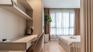 Moderni hotellihuone, jossa on sänky, työpöytä ja tuoli. Ikkunassa on verhot ja työpöydällä kasvi.
