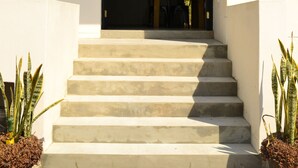 Ein Gebäude mit rotem Dach, beige farbenen Wänden und einer Treppe aus Beton, die zu einer dunklen Tür führt. An beiden Seiten der Treppe stehen je ein Blumentopf.