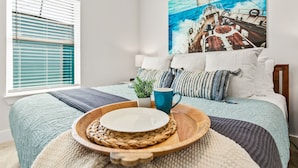 A neatly arranged bedroom with a bed, a window with blinds, a painting of a ship on the wall, and a tray with a plate and mug on the bed.