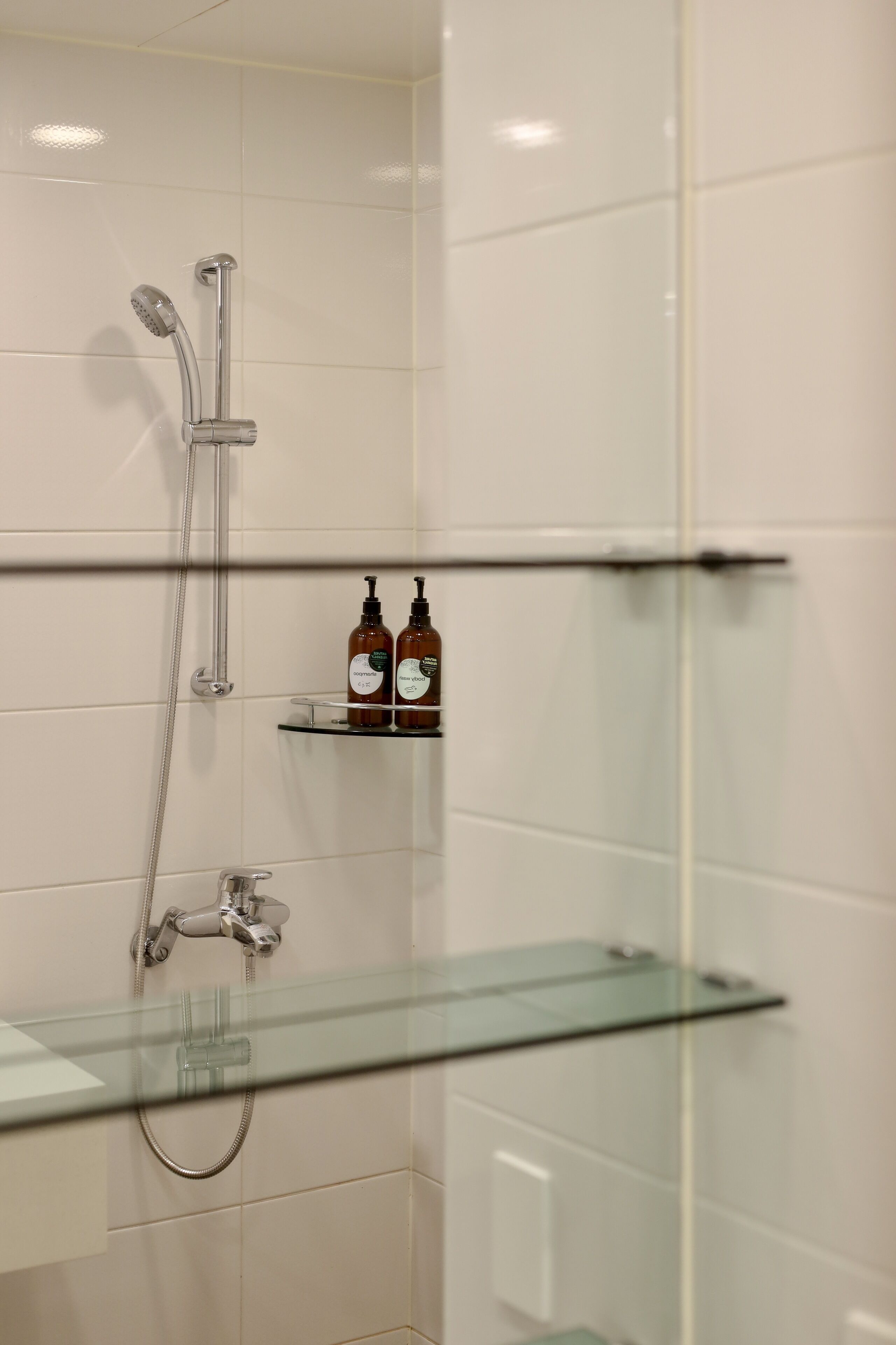 Une salle de bain avec des murs recouverts de carreaux blancs, une douche en verre et deux étagères remplies de bouteilles.