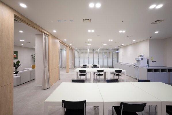 Une salle de conférence moderne, dotée de tables et de chaises blanches, d’un coin cuisine avec des appareils électroménagers et d’un espace détente avec un canapé et des plantes.