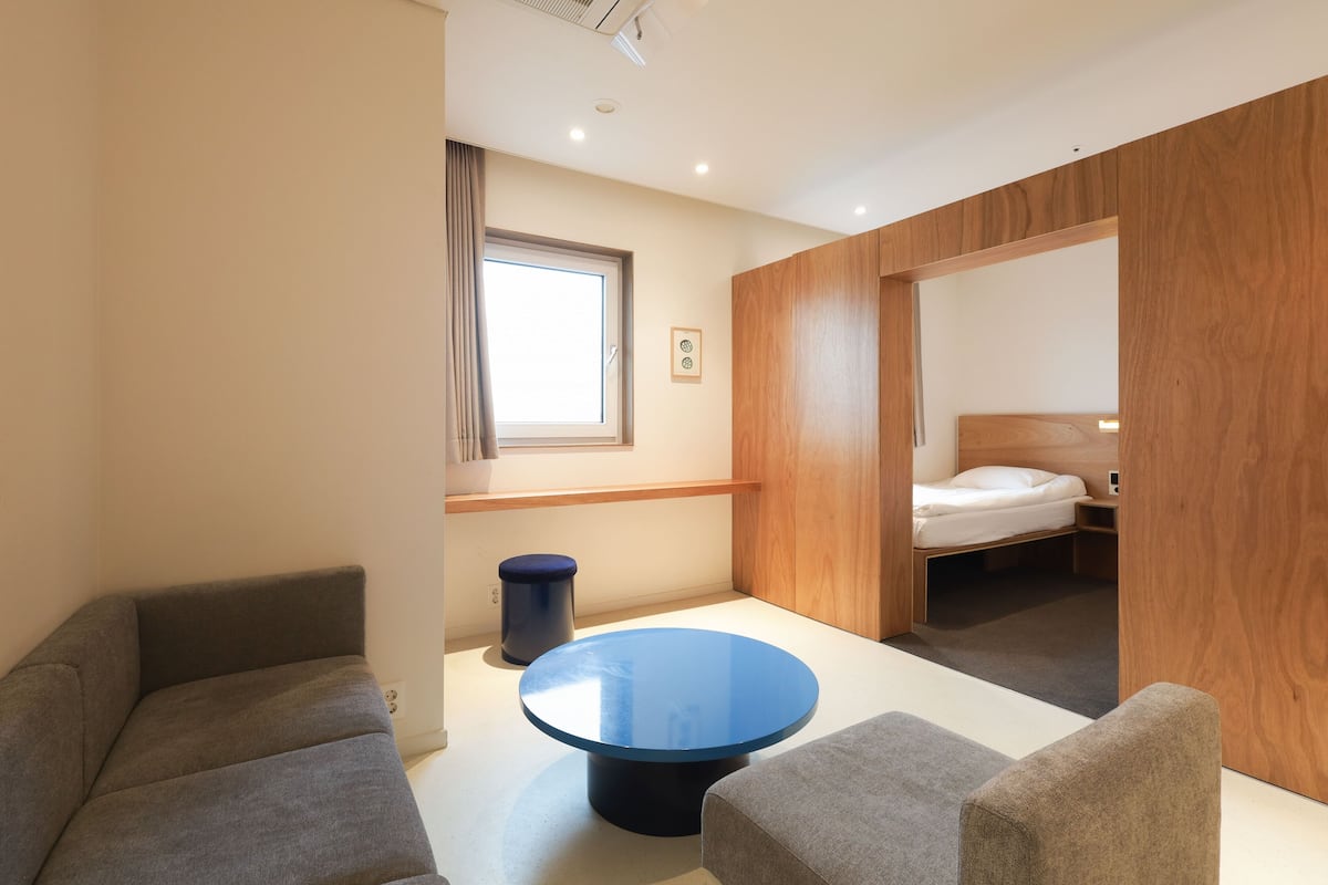 A modern hotel room with a round blue table, two grey sofas, a wooden wardrobe, a single bed, and a window with curtains.