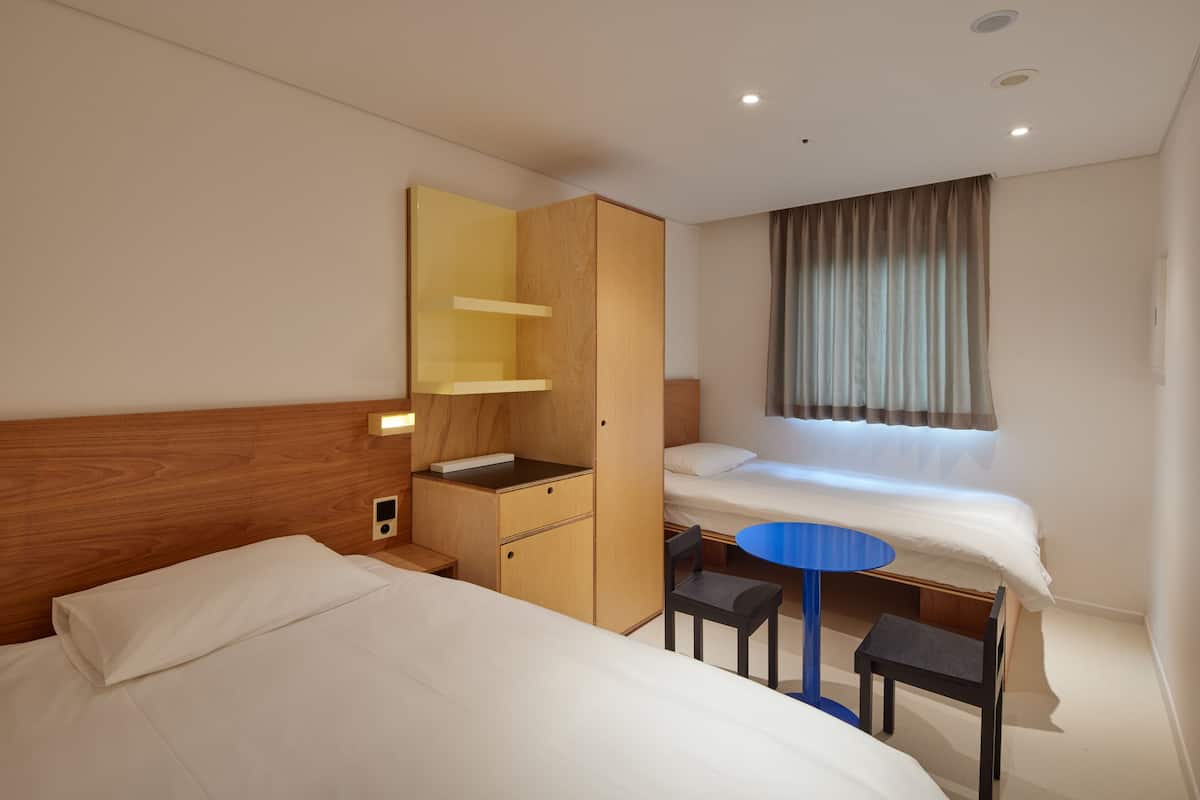 A hotel room with two beds, a wooden wardrobe, a small desk with a chair, and a round table.