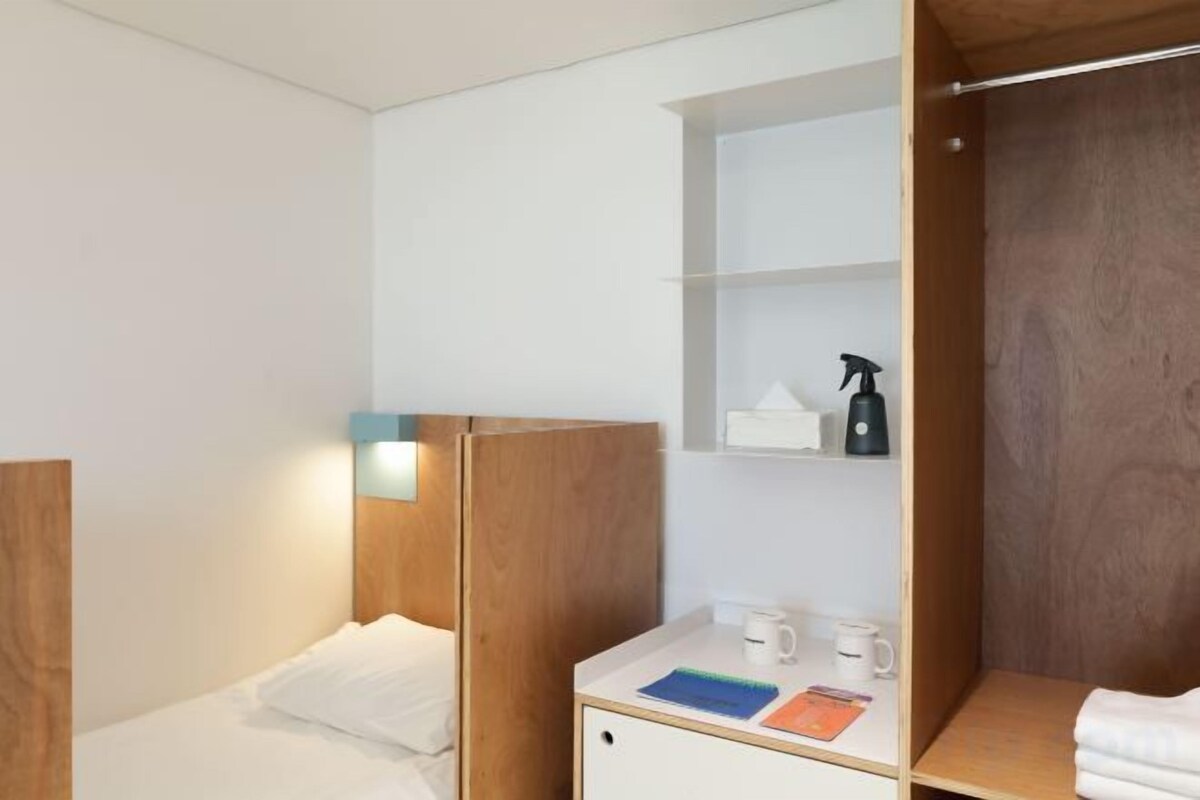 A hotel room with a bed, a bedside table with a lamp, a shelf with toiletries, and a wooden wardrobe.