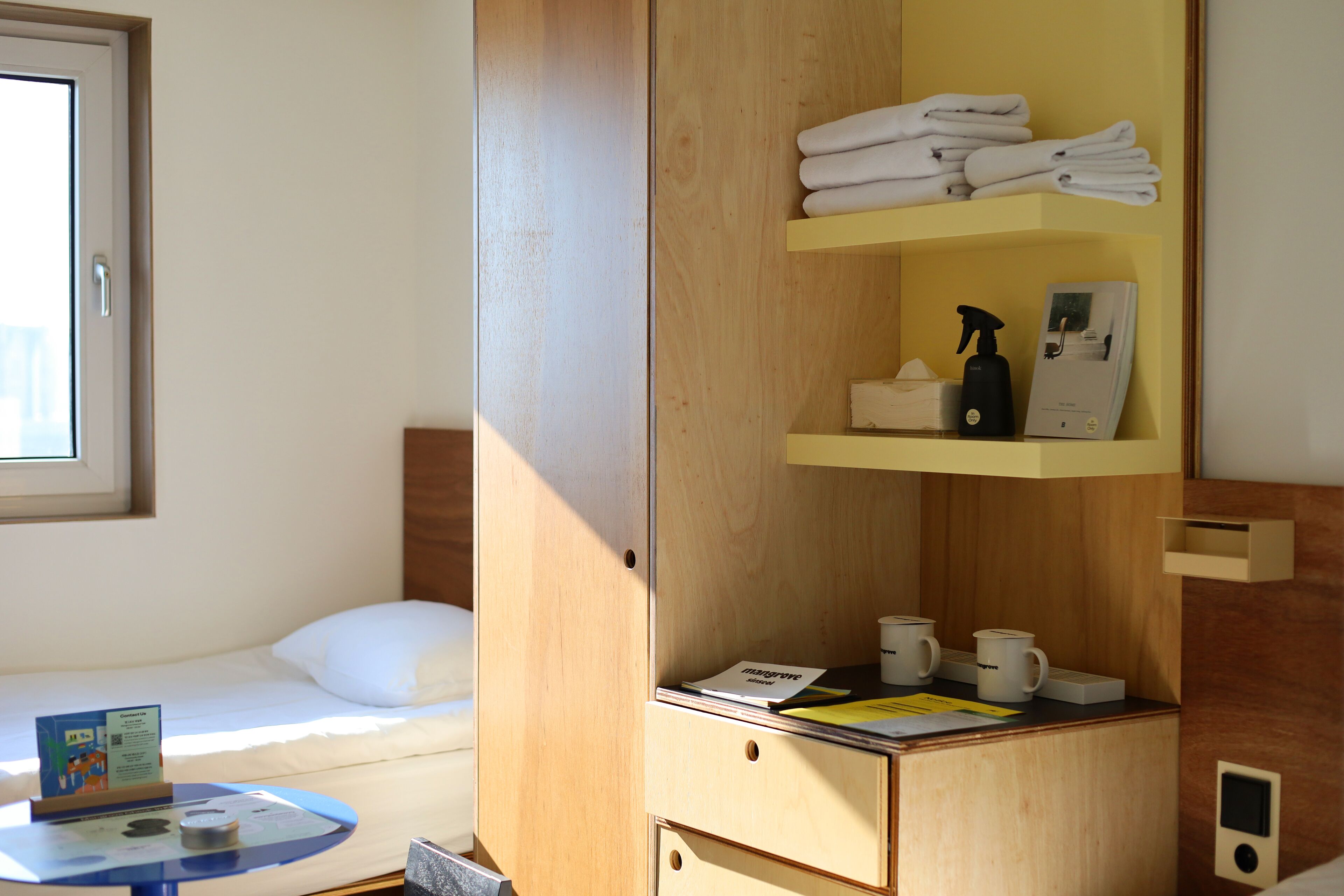 A hotel room with a bed, a small table with a book and a cup, a wooden wardrobe with towels and toiletries, and a window.