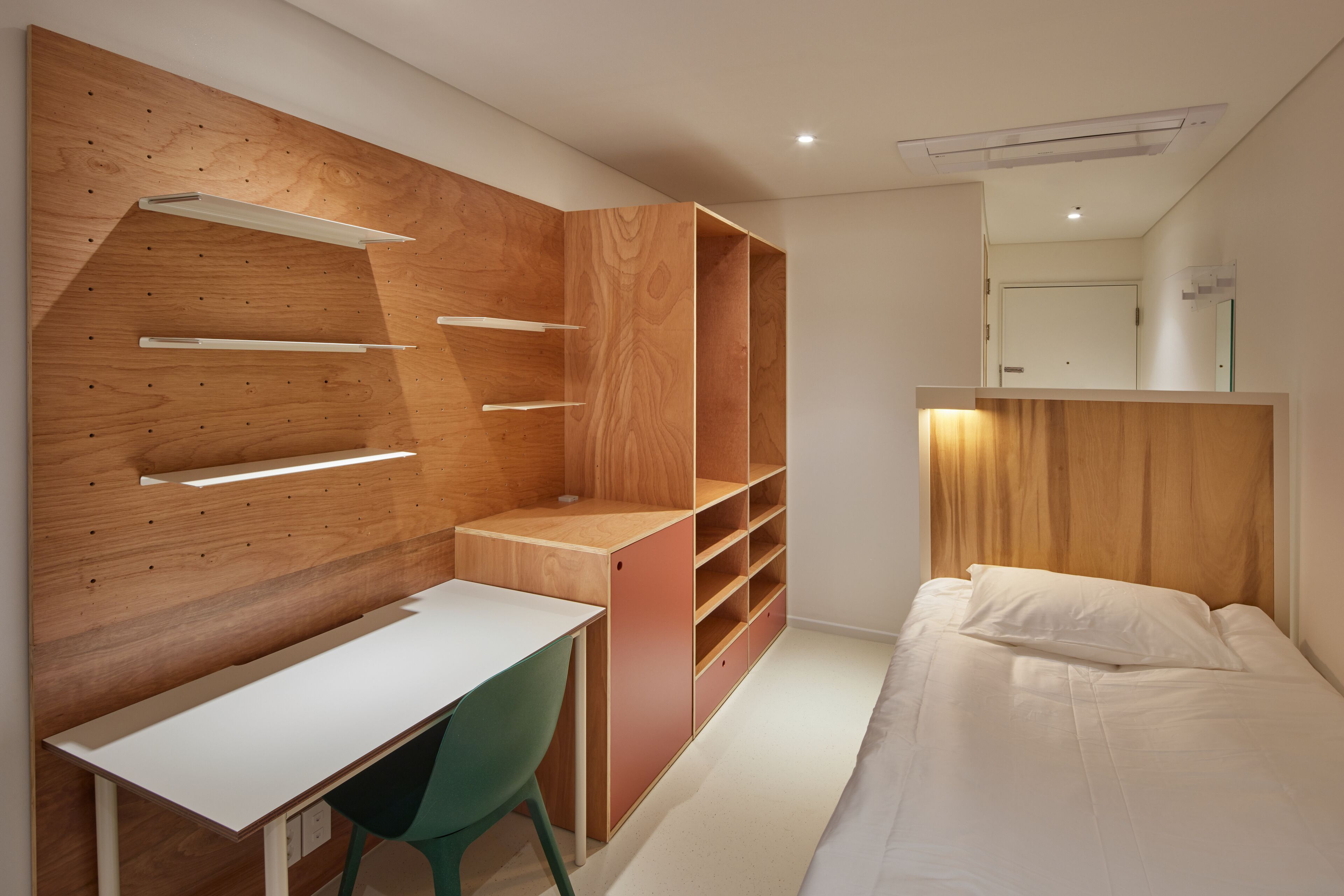 Ein modernes Schlafzimmer mit einer Holzwandverkleidung, einem weißen Schreibtisch, einem grünen Stuhl und einem Bett mit weißer Bettwäsche.