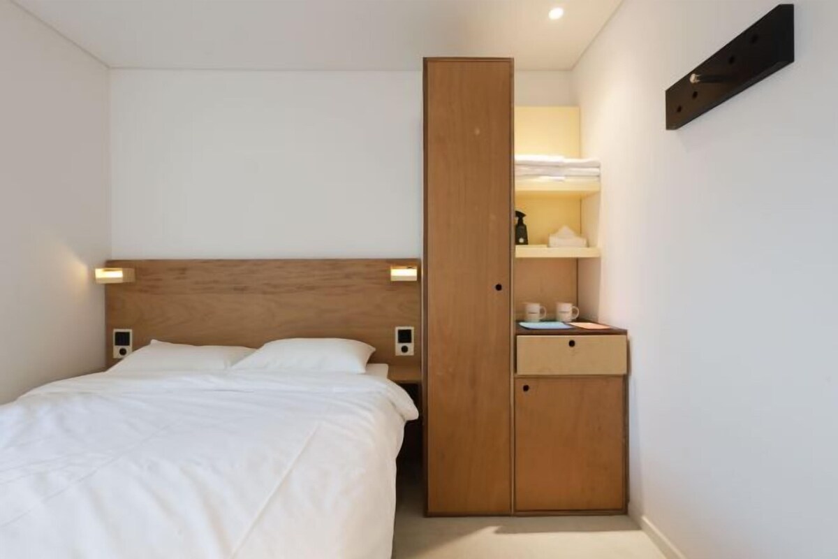 A bedroom with a bed, a wooden wardrobe, a bedside table with a lamp, and a shelf with towels.