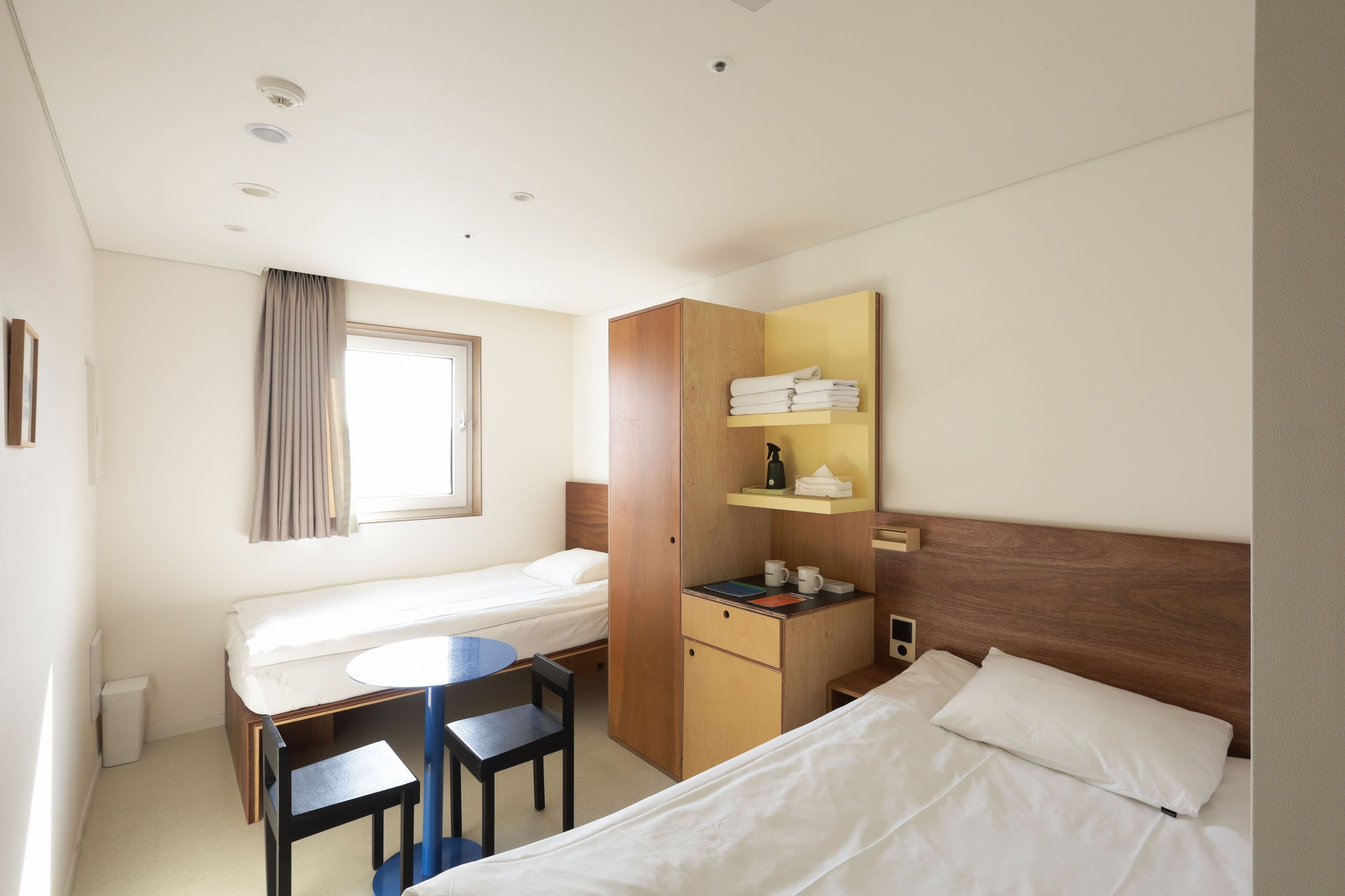 Habitación de hotel con dos camas individuales, un escritorio con silla, una mesita, un armario y un espejo.