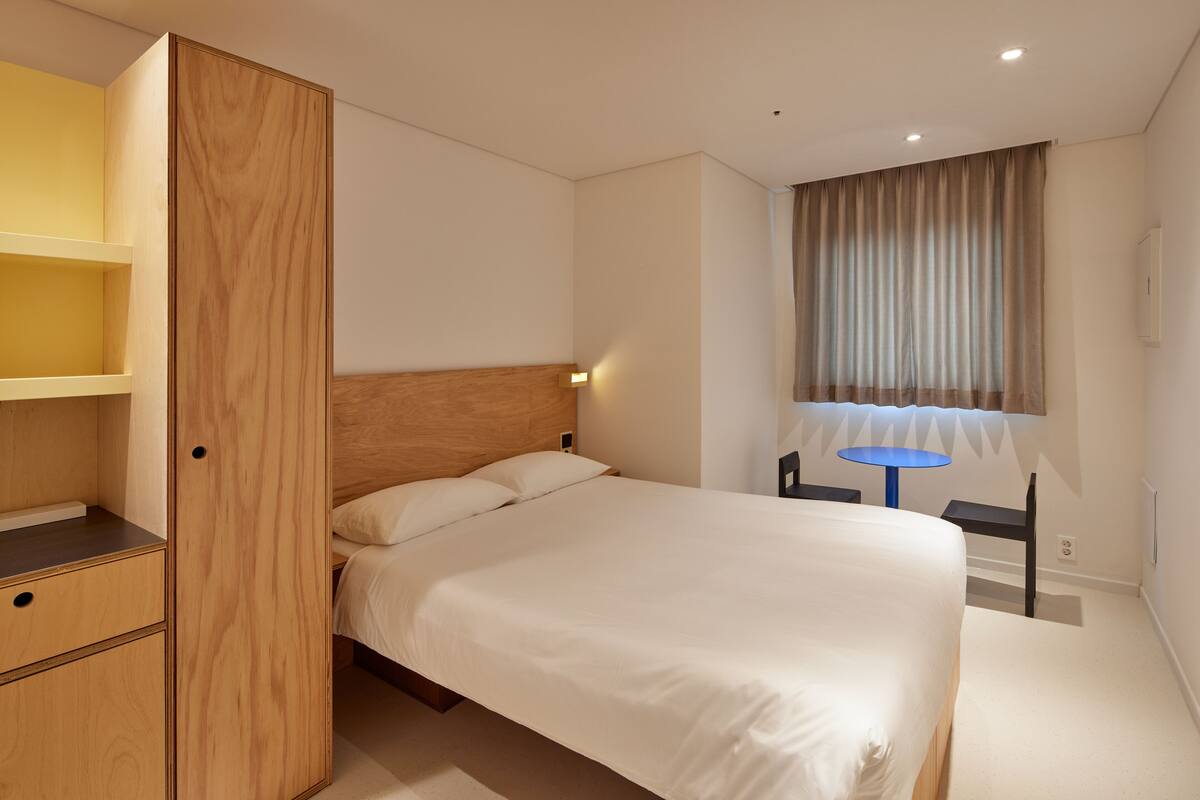 A hotel room with a bed, a wooden wardrobe, a desk, and a window with curtains.